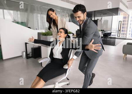 Colleagues playfully push smiling female coworker seated on office chair Stock Photo