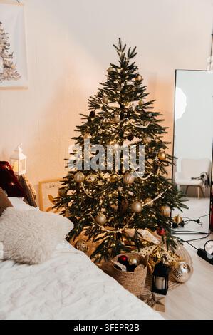 Tall fir tree decorated with gold baubles, pine cones and a wooden bead garland standing in a bright Scandinavian bedroom next to a large floor mirror Stock Photo