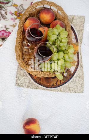 Wicker basket filled with ripe peaches and green grapes with two glasses of red wine on a painted wooden tray, standing on a white picnic blanket besi Stock Photo