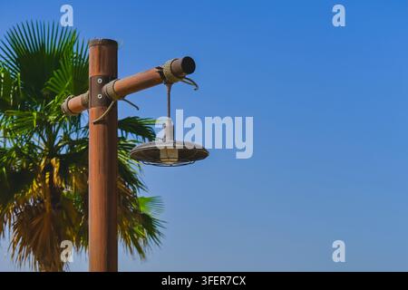 Vintage outdoor lamp hangs from wooden street lamppost beside palm tree against blue sky, rustic lighting with tropical eco-style. Stock Photo