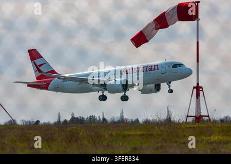 An Austrian Airlines Airbus A220 descends toward the runway with landing gear extended, a red-and-white windsock indicating wind direction. Stock Photo