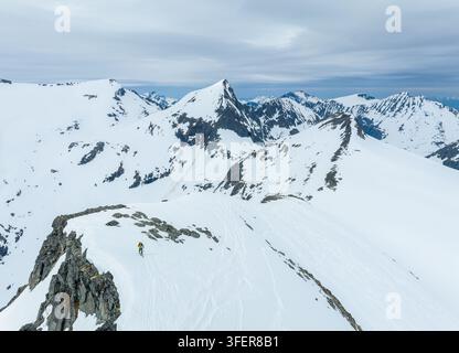 Aerial view of a lone skier traversing the snowy ridge of Ringshornet mountain surrounded by dramatic peaks under a cloudy sky in Fjord, More og Romsd Stock Photo