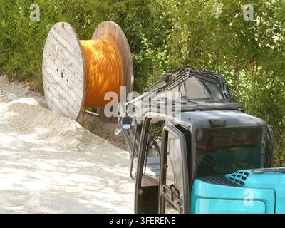 Aerial view of a road construction site featuring a large wooden cable spool with orange wiring and an excavator parked on gravel near green foliage i Stock Photo