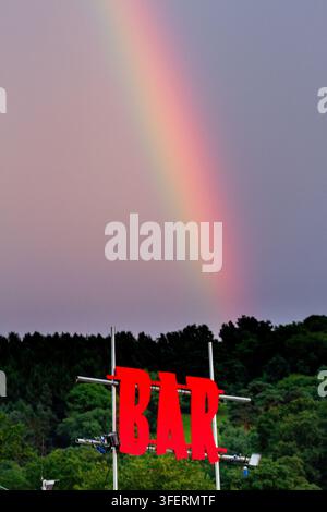 RAINBOW OVER THE FESTIVAL BAR, REPORTAGE, GREEN MAN FESTIVAL 2012, BRECON, WALES, UK, 2012: A vibrant rainbow arcs through a moody, overcast sky above a bright red bar sign and a line of dark green trees at an outdoor festival. Photo: Rob Watkins. INFO: The Green Man Festival is held annually in the Brecon Beacons in August. It celebrates alternative music, arts, literature and science in family-friendly mountain setting. The 2012 festival was the muddiest on record after a week of heavy rain. Stock Photo