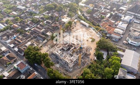 Aerial view of a construction site with two tall yellow cranes, concrete building frame, and surrounding residential neighborhood in Surakarta, Centra Stock Photo