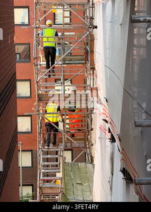 Three workers on multi level scaffolding between brick and sheet covered buildings. Stock Photo