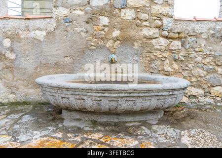 Ancient Stone Wall Fountain with Oval Basin and Brass Tap in Old European Village Street. Antique Open air bath tub with water. Backyard Stock Photo