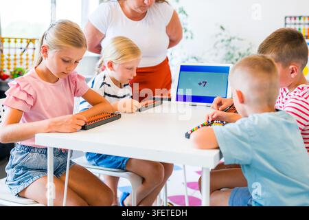 Teacher guides children practicing mental arithmetic and calculations with abacuses using a digital learning app during a classroom lesson. Stock Photo