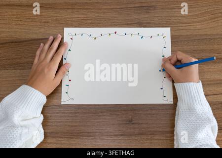 Hands sketching festive string lights border on blank sheet of paper on wooden table, blue pencil in use Stock Photo