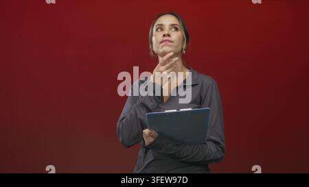 Woman holding a blue clipboard touches her chin in studio with red backdrop and casual sports top; thoughtful reflection. Stock Photo
