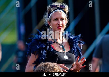 STYLISH WOMAN IN A FEATHER BOA AT A FESTIVAL, REPORTAGE, GREEN MAN FESTIVAL 2012, BRECON, WALES, UK, 2012: A stylish blonde woman wearing sunglasses on her head, a black top, and a blue feather boa around her shoulders smiles while gesturing with her hands. Photo: Rob Watkins. INFO: The Green Man Festival is held annually in the Brecon Beacons in August. It celebrates alternative music, arts, literature and science in family-friendly mountain setting. The 2012 festival was the muddiest on record after a week of heavy rain. Stock Photo