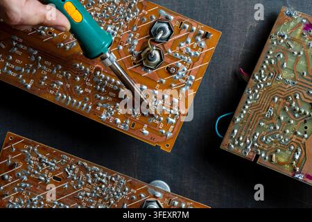 Close-up of a technician's hand soldering components on a vintage electronic circuit board with a green soldering iron. Ideal for repair services, eng Stock Photo