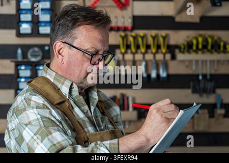 Senior male carpenter or craftsman taking notes in workshop. Elderly technician or carpenter wearing glasses and an apron writes in notebook against b Stock Photo