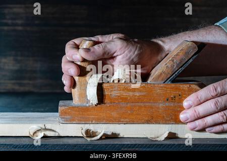 Craftsman hands using vintage wooden hand plane tool, close up. Carpenter hands smoothing timber plank with traditional wooden hand plane, creating sh Stock Photo