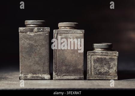 Three vintage rusty metal containers, close up. Antique industrial tin cans with screw caps covered in heavy rust and patina standing on dark surface Stock Photo