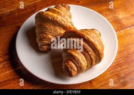 Two freshly baked, crispy and flaky golden croissants served on a clean white ceramic plate, placed on a rustic wooden table surface. Stock Photo