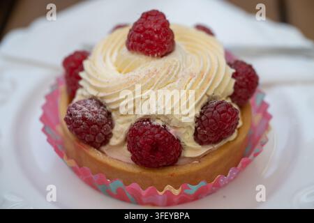 A close-up view of a delicate raspberry tartlet featuring a large swirl of whipped cream and fresh berries, served in a colorful paper liner on a deco Stock Photo