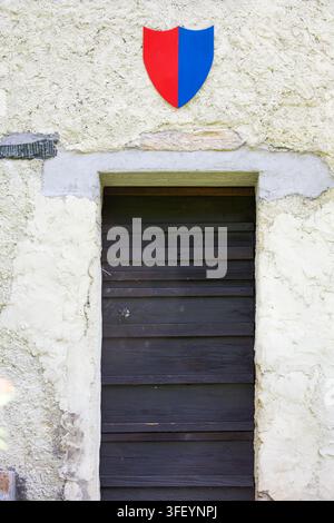 Red and blue coat of arms of Canton Ticino mounted above an ancient rustic wooden entrance door on a stone building wall in Ticino, Switzerland. Stock Photo