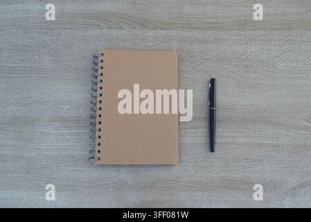 Top-down vertical flat lay composition showing a closed brown kraft spiral notebook paired with a professional black pen, centered perfectly on a clea Stock Photo