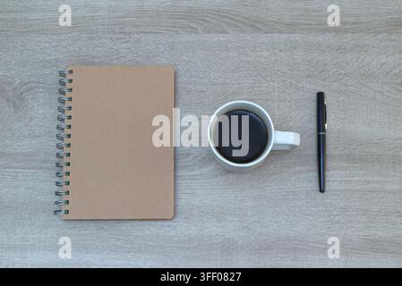 Top-down flat lay composition featuring a spiral kraft notebook, a white ceramic coffee mug, and a black pen aligned on a light wooden desk surface, s Stock Photo