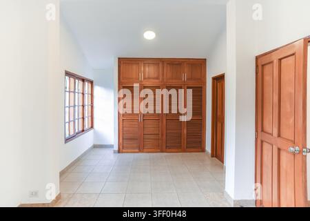 Spacious Room with Wooden Wardrobe and Window: An empty, well-lit room featuring a large built-in wooden wardrobe, tiled floor, and a window Stock Photo