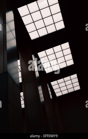 A dramatic upward perspective captures three rectangular luminous ceiling panels divided into square grids, suspended in deep shadow above tall vertic Stock Photo