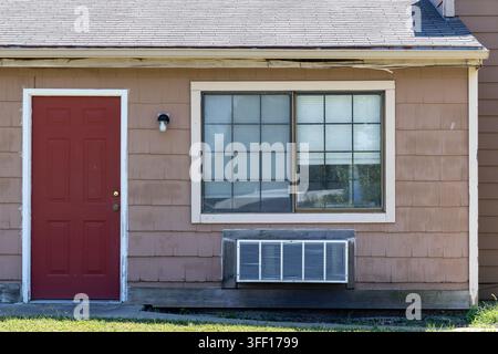 Front exterior of an older brown shingle-sided residence with a red door, large window and through-wall air-conditioning unit beneath visibly weathere Stock Photo