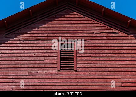 xCropped architectural view of the gable end of a weathered red wooden barn with horizontal siding, central louvered vent and deep geometric shadows b Stock Photo