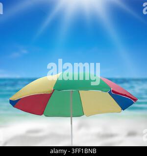 A vibrant multi-colored parasol stands on a sandy beach against a blurred tropical ocean and bright sunny sky Stock Photo