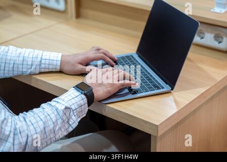 Close-up of a man wearing a smartwatch and checkered shirt typing on a modern laptop computer keyboard at a wooden office desk, symbolizing remote wor Stock Photo