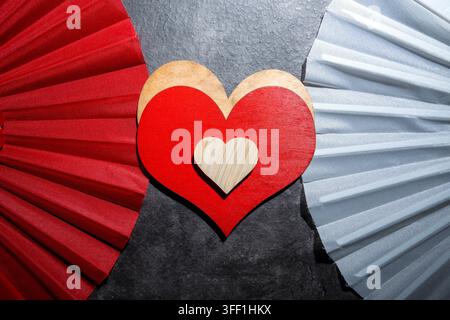 Artistic composition featuring nested heart shapes in red and wood, positioned between decorative red and silver paper fans against a dark textured ba Stock Photo