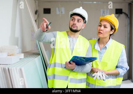 Caucasian man and asian woman builders discussing project documentation in apartment. Pointing finger gesture. Stock Photo