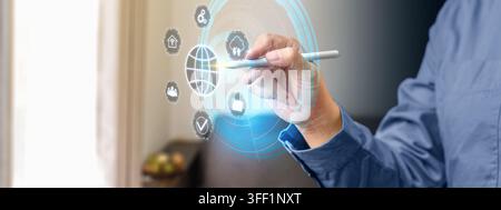 A man in a blue shirt uses a stylus to select icons on a glowing futuristic digital HUD interface displaying global connection, security, and smart ho Stock Photo