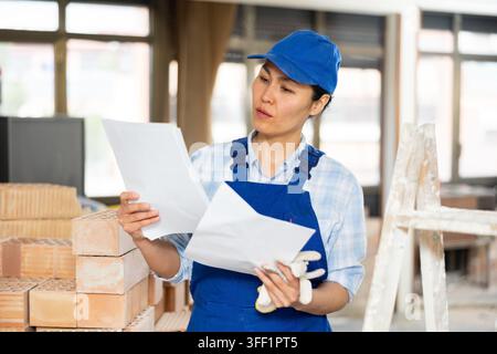 Focused woman architect wearing uniform examining indoor building construction site and making notes for workmen Stock Photo