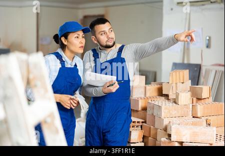 Caucasian man and asian woman builders discussing project documentation in apartment. Pointing finger gesture. Stock Photo