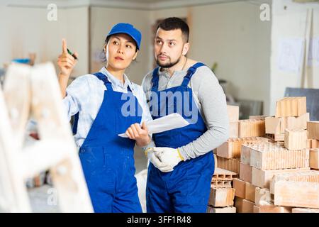 Professional male and female contractors workmates in blue uniform standing with papers in hands and discussing in building during renovation Stock Photo