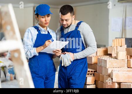 Professional male and female contractors workmates in blue uniform standing with papers in hands and discussing in building during renovation Stock Photo