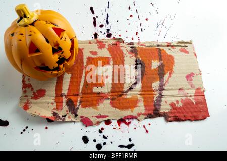 Top-down view of jack-o'-lantern pumpkin sitting on piece of cardboard with the word HELP painted on it, splattered with blood on a white background, Stock Photo