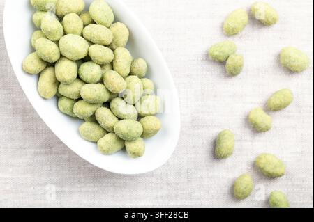 Wasabi peanuts in a white bowl, on linen. Crunchy and spicy snack made of peanut kernels, coated in savory, green wasabi-flavored shell. Photo Stock Photo