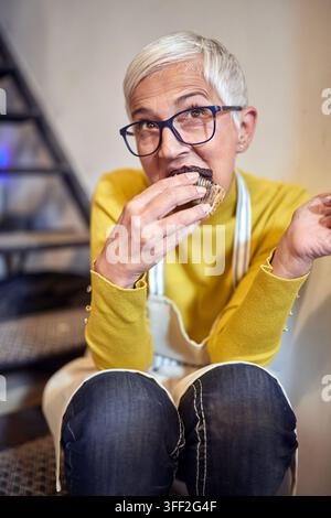 happy senior woman taking a break eating cupcake after making them in confectionery shop Stock Photo