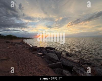 Serene coastal sunset scene with sandy beach and rocky shoreline under a dramatic sky with scattered clouds and a distant tree line on the horizon. Stock Photo