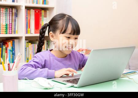 Smiling elementary schoolgirl typing on a laptop computer while studying at home desk. Stock Photo