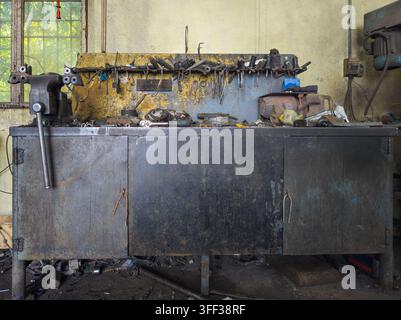 Cluttered metal workbench in workshop features heavy duty vice and various tools. Visible mechanical parts and a window with greenery enhance the indu Stock Photo