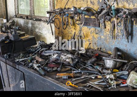 Cluttered workbench filled with various tools and mechanical parts in a busy workshop. A robust vise is prominently displayed on the left side, showcasing a functional repair environment. Stock Photo