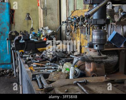 Cluttered workbench filled with tools and machinery in a busy workshop. Drill press and heavy-duty vise dominate the scene, showcasing an active repair environment. Stock Photo
