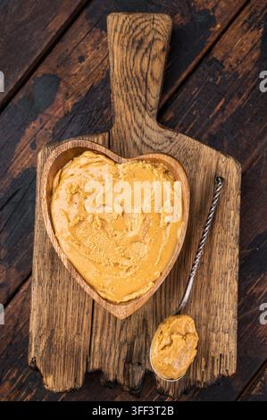Creamy peanut butter spread in heart shaped wooden bowl on rustic cutting board with spoon. Natural peanut paste made from roasted peanuts Stock Photo