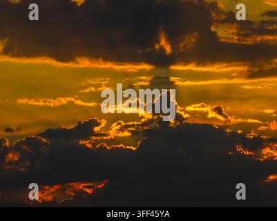 Dramatic sunset sky features dark clouds silhouetted against a vibrant golden-orange backdrop. Rays of sunlight pierce through, creating an awe-inspiring atmosphere. Stock Photo