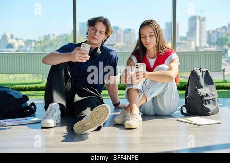 Male and female college students sitting on floor and using their smartphones independently. Young couple or classmates absorbed in mobile phones near Stock Photo
