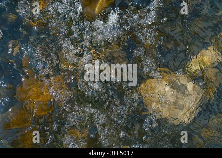 Close-up view of clear, moving water with sparkling bubbles flowing over smooth river rocks. Stock Photo