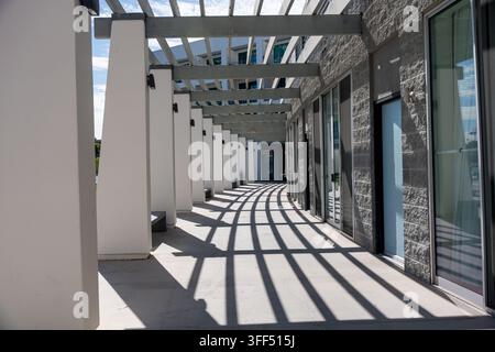 A modern outdoor pedestrian walkway features repeating columns, overhead beams, glass doors, and dramatic geometric shadows Stock Photo